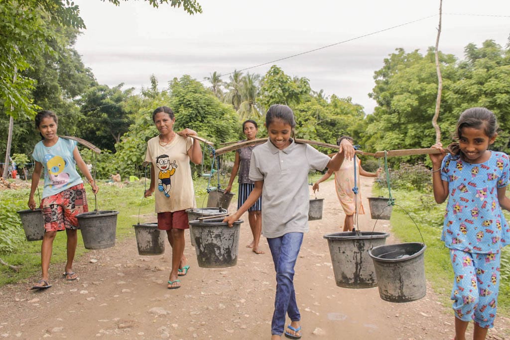 A group of women and girls are carrying water in buckets hanging from yokes on their shoulders. They are walking down a dirt road.